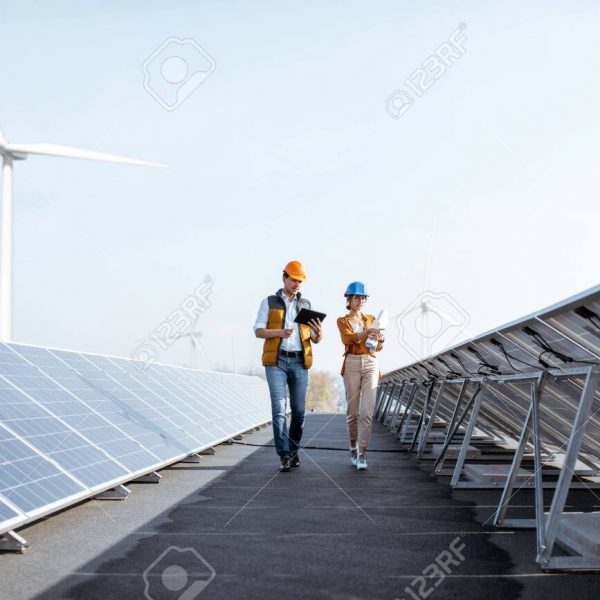 View on the rooftop solar power plant with two engineers walking and examining photovoltaic panels. Concept of alternative energy and its service